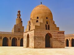 Ibn Khaldun’s Maghreban and Al-Andalusian Journey: A New Biography Explored Ibn Khaldun sitting at a desk, writing with a quill pen, surrounded by books and maps of Al-Andalusia and the Maghreb